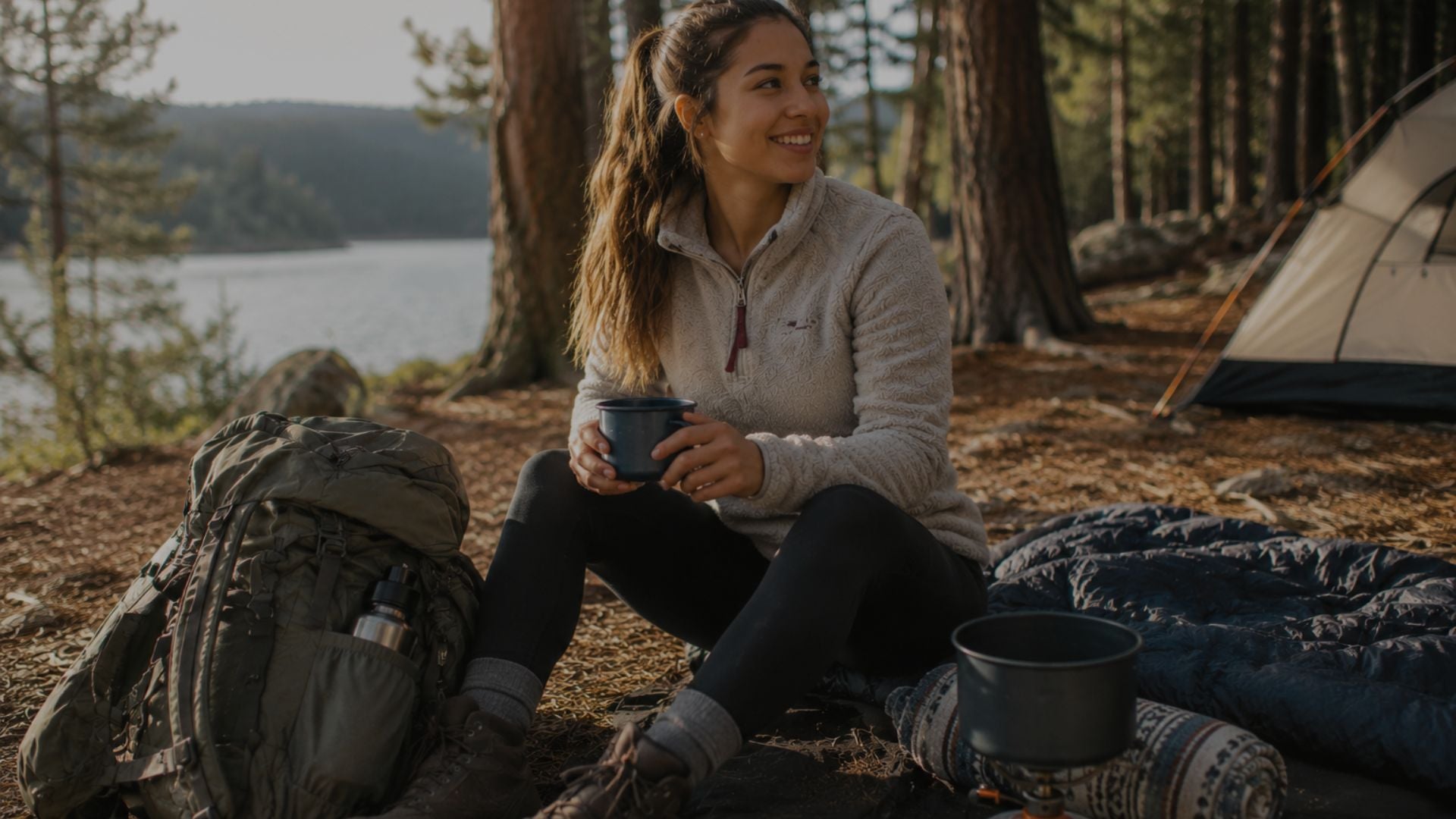 Woman sitting in a forested area with a lake, holding a mug, next to a backpack and camping gear.